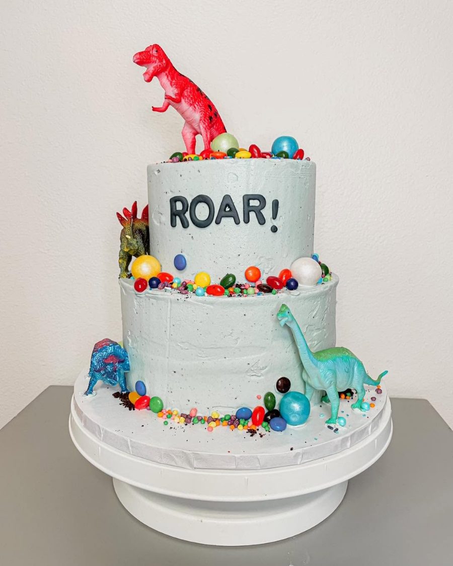 Cakes & Bakes's own photograph: a two tier pale blue dinosaur cake reading ROAR, with toy dinosaurs and candy.