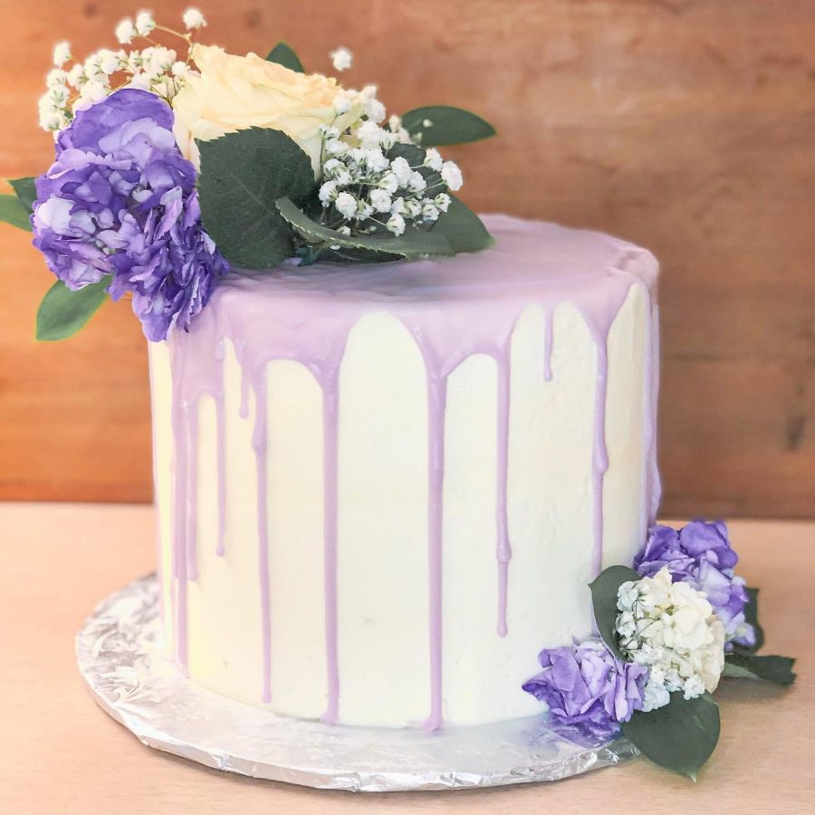 Cakes & Bakes's own photograph: a lavender and white drip cake dressed with fresh purple florals.